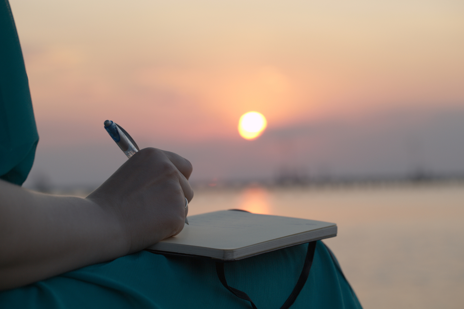 Close up view of the hand of a woman writing in her diary at sunset with the glowing orb of the sun reflected over a still ocean